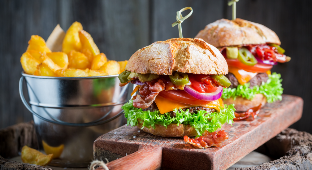Zwei Burger mit Bacon, Käse, Salat und Gemüse auf Holzbrett, daneben Eimer mit knusprigen Pommes in rustikalem Stil
