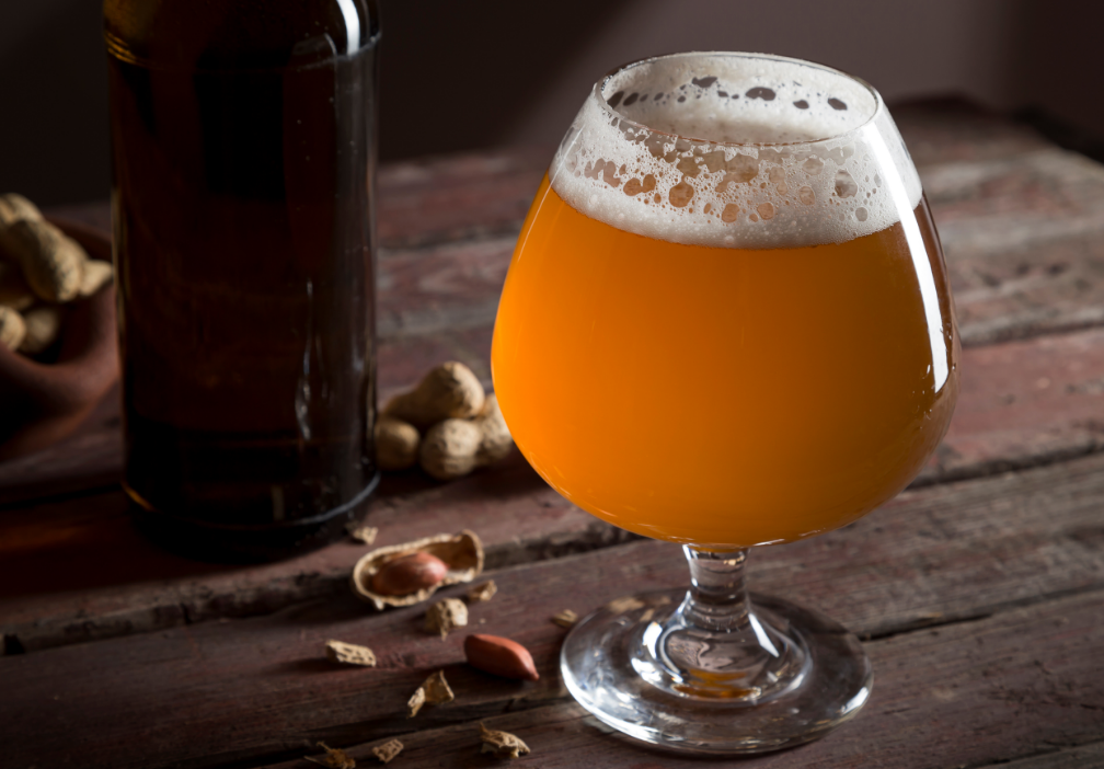 Pale Ale in bauchigem Glas auf rustikalem Holztisch, daneben Erdnussschalen und eine dunkle Bierflasche im Hintergrund